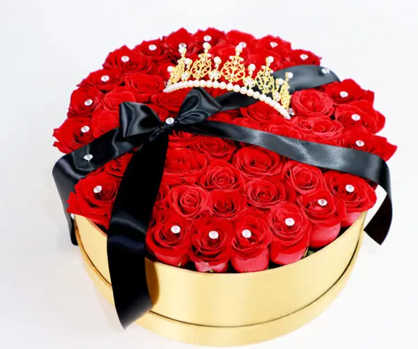 Caja dorada con rosas rojas frescas, decorada con brillantes y una corona dorada con perlas, complementada con un lazo negro