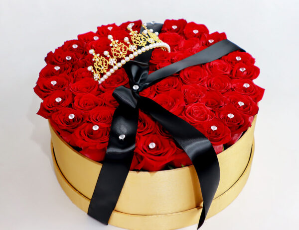 Caja dorada con rosas rojas frescas, decorada con brillantes y una corona dorada con perlas, complementada con un lazo negro