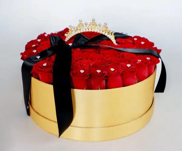 Caja dorada con rosas rojas frescas, decorada con brillantes y una corona dorada con perlas, complementada con un lazo negro